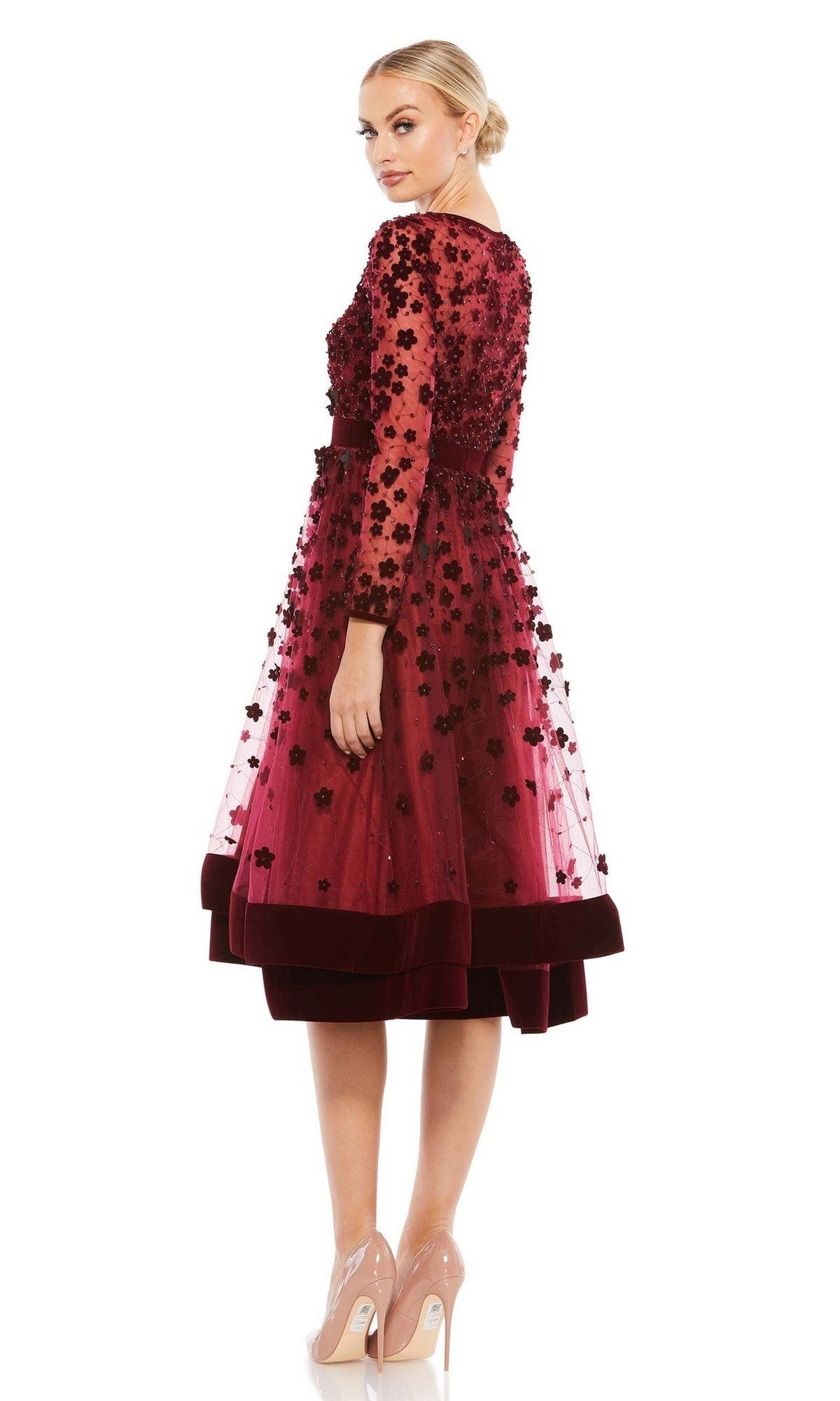 A woman models the Mac Duggal 67007, a knee-length wedding-guest dress with floral embellishments, sheer overlay, and long sleeves, paired with nude heels against a white background.